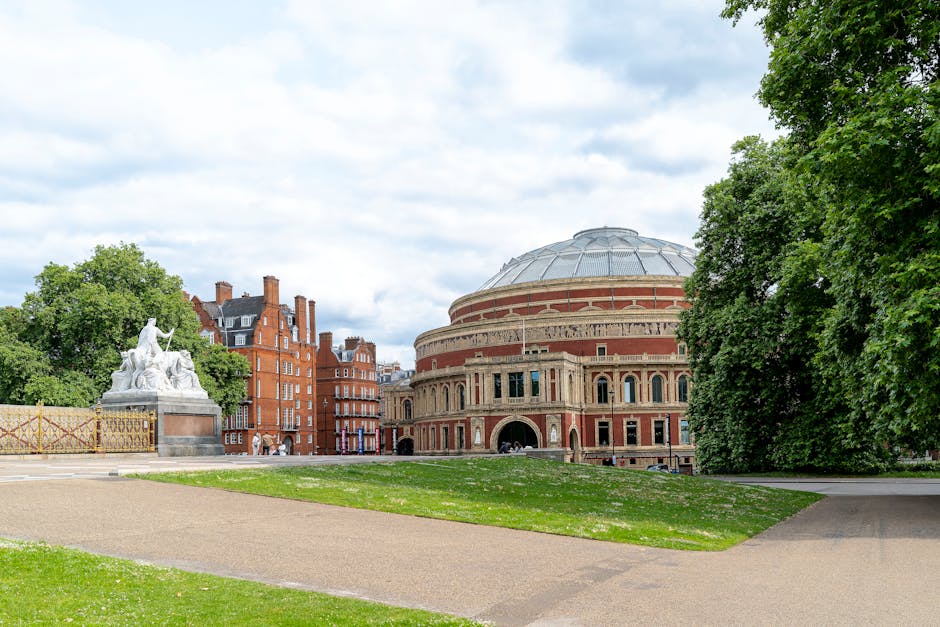 A wide-angle view of a house removal scene in Kensington showing the loading process outside a large, historic building with a domed roof and ornate brickwork. In the foreground, a gravel pathway curves through well-maintained grassy areas, with a green hedge on the right side. On the left, a white stone sculpture of seated figures is partially visible, surrounded by a decorative metal fence. Visible inside the property, there are cardboard moving boxes and bubble wrap, indicating packing materials used during home relocation. A Man with Van Kensington vehicle is parked nearby, with its side door open, ready for furniture transport. The scene captures the outdoor environment with soft natural lighting under a partly cloudy sky, emphasizing a professional and organized moving process involving careful packing, loading, and transportation of household items in an urban setting.