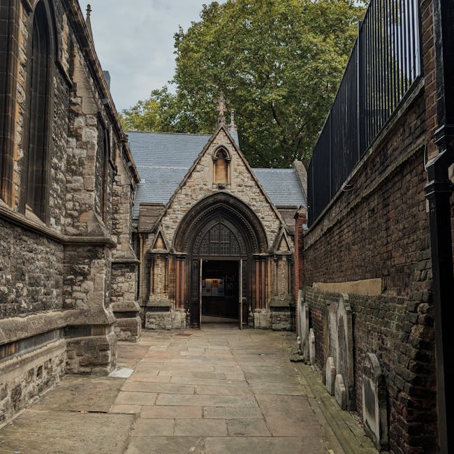 A narrow stone-paved pathway leads to the entrance of a historic church with Gothic architectural features, including an arched doorway and intricate stone detailing, set between two brick walls. The church's facade is constructed of weathered stone, with a pointed arch window above the entrance. To the right, a tall brick wall topped with a black metal fence runs alongside the pathway. On the left, part of the church's stone wall is visible, with a large stained glass window partially obscured. The pathway appears to be inside a historical ecclesiastical site, with trees visible in the background under a cloudy sky. The scene captures an outdoor setting, likely during daytime, with natural light illuminating the stone and brick materials. This image exemplifies the structural complexity of historic buildings, relevant to home relocation or moving services dealing with specialty preservation of valuable or delicate architecture, provided by Man with Van Kensington.