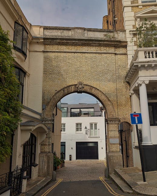 A wide-angle view of a house removal scene in Kensington showing the loading process outside a large, historic building with a domed roof and ornate brickwork. In the foreground, a gravel pathway curves through well-maintained grassy areas, with a green hedge on the right side. On the left, a white stone sculpture of seated figures is partially visible, surrounded by a decorative metal fence. Visible inside the property, there are cardboard moving boxes and bubble wrap, indicating packing materials used during home relocation. A Man with Van Kensington vehicle is parked nearby, with its side door open, ready for furniture transport. The scene captures the outdoor environment with soft natural lighting under a partly cloudy sky, emphasizing a professional and organized moving process involving careful packing, loading, and transportation of household items in an urban setting.