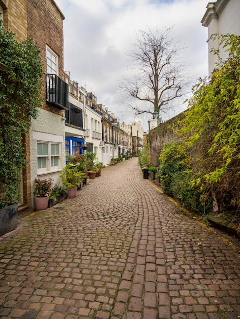 A narrow cobblestone street in Kensington, lined with terraced houses featuring white facades, black wrought-iron balconies, and large front windows. Potted plants and small shrubs are placed along the sidewalk, and a tall, leafless tree is visible in the background under a cloudy sky. The scene depicts a quiet residential area possibly involved in home relocation or furniture transport, with no visible moving equipment or vehicles in this image but consistent with the context of house removals and packing and moving services provided by Man with Van Kensington.