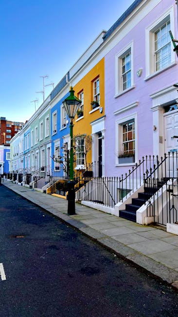 A narrow cobblestone street in Kensington, lined with terraced houses featuring white facades, black wrought-iron balconies, and large front windows. Potted plants and small shrubs are placed along the sidewalk, and a tall, leafless tree is visible in the background under a cloudy sky. The scene depicts a quiet residential area possibly involved in home relocation or furniture transport, with no visible moving equipment or vehicles in this image but consistent with the context of house removals and packing and moving services provided by Man with Van Kensington.