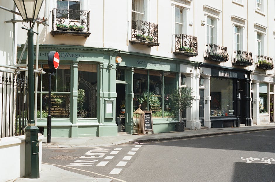 A street scene outside a small retail shop with a light green facade and large front windows displaying floral arrangements, positioned at the corner of a narrow street. The shop has decorative elements with a black signboard and a chalkboard menu placed on the sidewalk in front. Adjacent to the shop is a white building with tall windows and small balconies with black wrought iron railings, some adorned with potted plants. A bicycle lane with white bike symbols is painted on the asphalt, and there's a no-entry traffic sign attached to a pole near the shop entrance. In the foreground, a black bollard and a metal pole are visible, while a metal fencing runs along the street edge. This scene illustrates an urban environment suitable for home relocation activities, with considerations for narrow access streets and the need for careful furniture transport, as often managed by companies like Man with Van Kensington during removals.