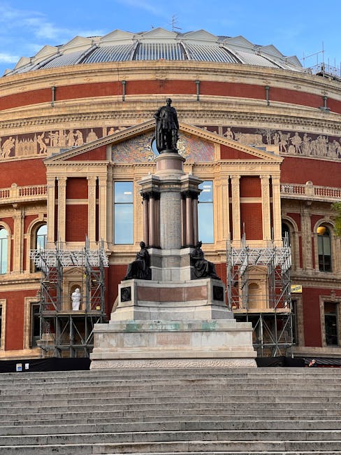 A wide view of the Royal Albert Hall in Kensington during daytime, with its distinctive domed roof and ornate brick and stone façade visible. In front of the building, there is a large stone statue of a standing figure mounted on a tall pedestal, flanked by seated statues at its base. Scaffolding surrounds parts of the entrance area, indicating ongoing maintenance or restoration work. The scene is outdoors, with steps leading up to the hall’s entrance and a clear blue sky overhead. This setting is relevant for house removals and furniture transport, highlighting the architectural context often involved in moving logistics and home relocation projects, as coordinated by services like Man with Van Kensington.