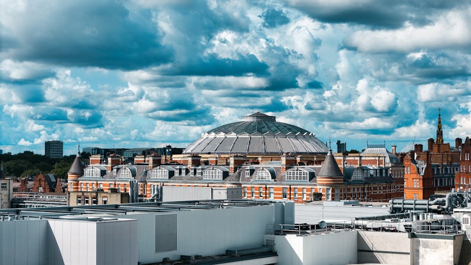 A wide view of the Royal Albert Hall in Kensington during daytime, with its distinctive domed roof and ornate brick and stone façade visible. In front of the building, there is a large stone statue of a standing figure mounted on a tall pedestal, flanked by seated statues at its base. Scaffolding surrounds parts of the entrance area, indicating ongoing maintenance or restoration work. The scene is outdoors, with steps leading up to the hall’s entrance and a clear blue sky overhead. This setting is relevant for house removals and furniture transport, highlighting the architectural context often involved in moving logistics and home relocation projects, as coordinated by services like Man with Van Kensington.