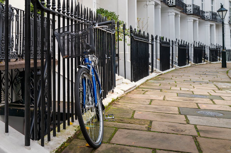A blue bicycle with a front-mounted black wire basket is locked to black metal railing outside a row of white terraced houses in Kensington, London. The bicycle is positioned next to a white staircase leading to a front door, with the bike's rear wheel resting on a paved sidewalk made of large, rectangular stone slabs. The pavement shows some moss growing between the stones. The black metal railings, featuring pointed finials, extend along the length of the property line, separating the houses from the street. In the background, the façade of the houses includes white columns, black wrought iron balconies, and traditional street lamps mounted on the exterior walls, indicating a classic London residential area. The image captures a typical scene during a home relocation or moving process, where furniture or boxes might be carried outside, and vehicles such as Man with Van Kensington are often involved in transporting belongings. The natural daylight and shadow indicate a clear, sunny day, ideal for moving activities.
