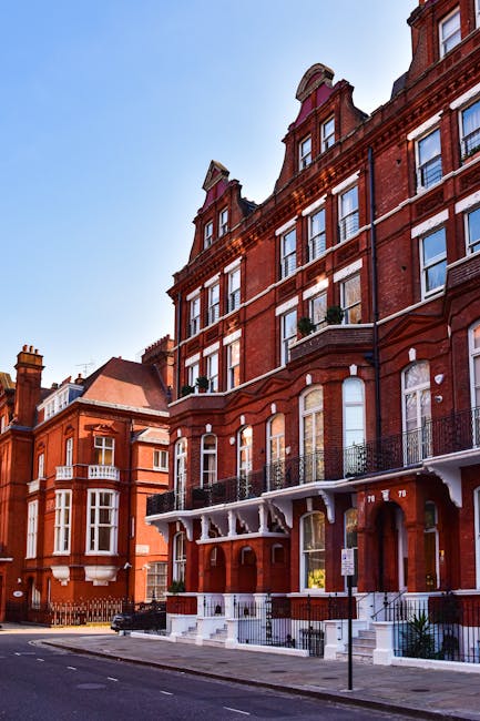 A quiet residential street in Kensington lined with parked cars on both sides, including compact and mid-sized vehicles in various colors such as black, white, blue, and grey. Elegant Victorian-style terraced houses with detailed facades and large windows run along the pavement, some with small front gardens or steps leading to entry doors. Blooming pink blossom trees with dense clusters of flowers extend their branches over the street, creating a vibrant canopy against the partly cloudy sky. In the foreground, a man with a van, associated with Man with Van Kensington, is visible preparing for a home relocation or furniture transport, with a load of cardboard boxes and wrapped furniture visible near the vehicle, suggesting an ongoing moving process. The scene depicts typical residential moving logistics, including loading or unloading activities in a peaceful neighbourhood setting, with natural daylight illuminating the area clearly.