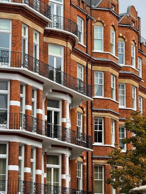 A row of white residential buildings with black wrought iron railings along a paved sidewalk in Kensington, London. A blue bicycle is parked against one of the railings, and climbing plants with pink