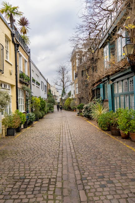 A cobbled residential street in South Kensington, London, with multi-storey houses on either side. The buildings feature a mix of brick and painted facades, with some having flower boxes and potted plants outside. Leafless trees line the street, indicating a winter season. In the distance, two pedestrians walk along the pavement. The scene is well-lit with natural daylight, and the overall atmosphere is calm and quiet, typical of an area suitable for house removals and home relocation activities. The street's narrow width and vintage character highlight the importance of professional removals services like those provided by Man with Van Kensington to facilitate efficient furniture transport and packing during local moves.