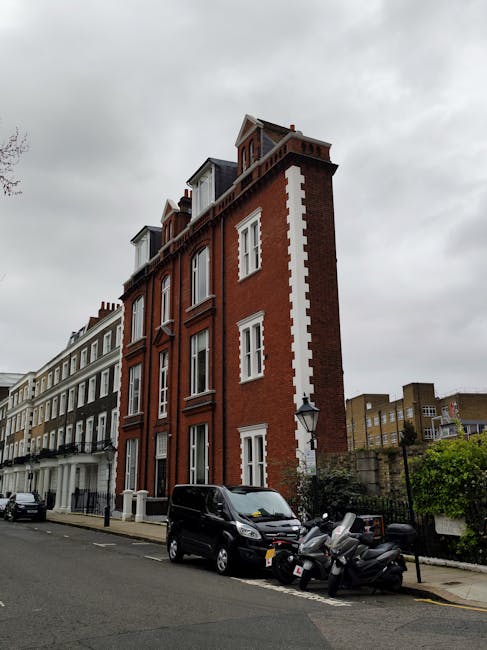 A multi-storey red brick building with white window frames and decorative elements, situated on a residential street in Kensington. The building features a narrow, vertical structure with dormer windows on the upper floors and a small rooftop extension. Parked along the pavement in front are a black van and several scooters, indicating a busy, urban setting. The street is lined with similar terraced houses, some painted white, with a cloudy sky overhead. The scene reflects a typical home relocation environment, where nearby vehicles suggest moving or furniture transport activities, consistent with professional removals services in the Kensington area, such as those provided by Man with Van Kensington.